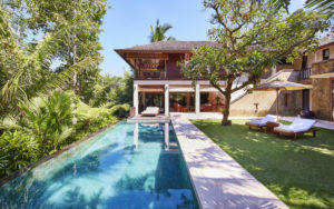 Daytime view of the infinity pool surrounded by the Bali jungle at the Pita Linggar 3 bedroom home with sun loungers and a parasol adjacent on the grass lawn