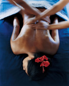 Female with a flower in her hair lying on a treatment bed receiving a massage from two people